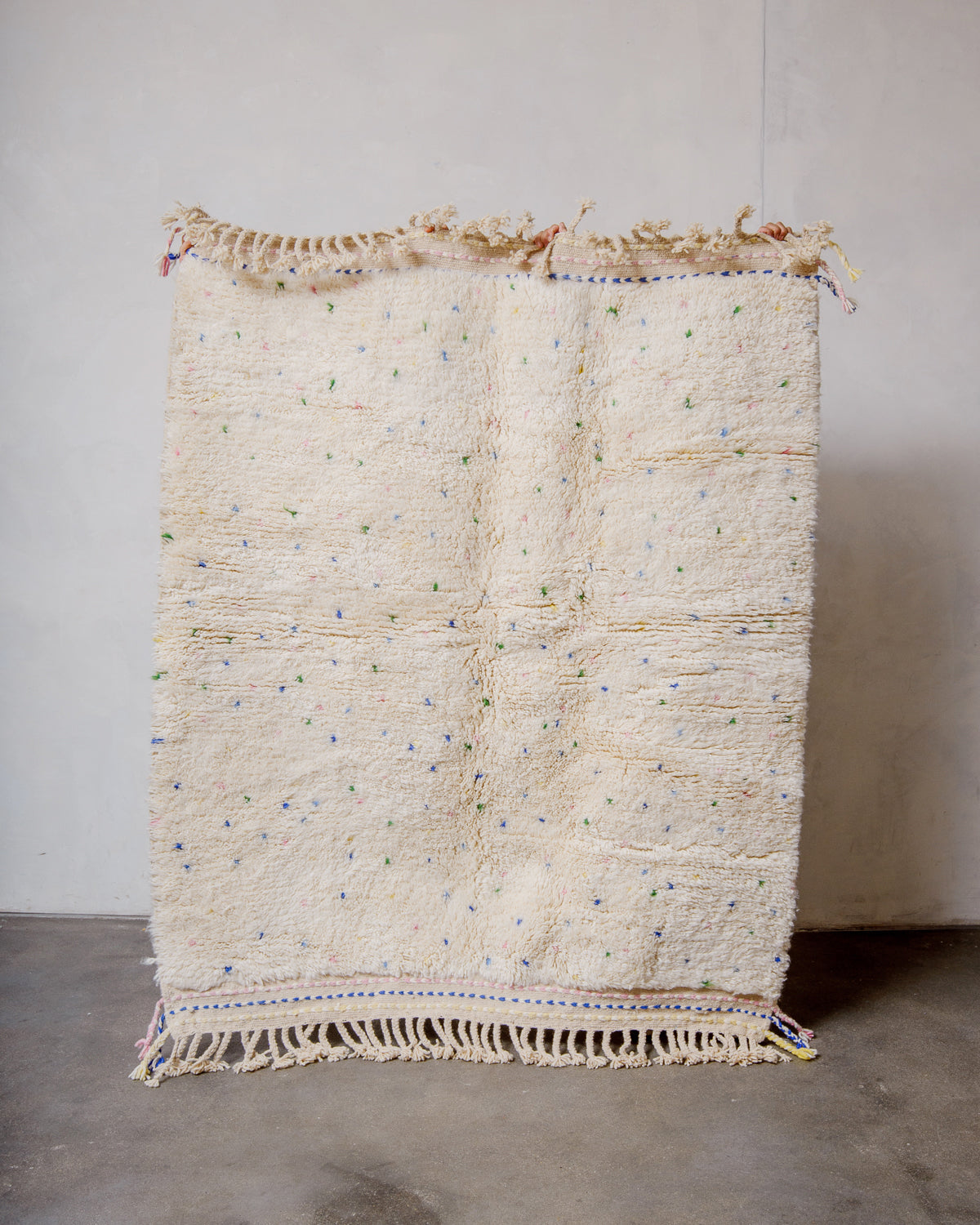 Berberlin Beniourain rug in cream wool with coloured dots being held up in front of a white wall.